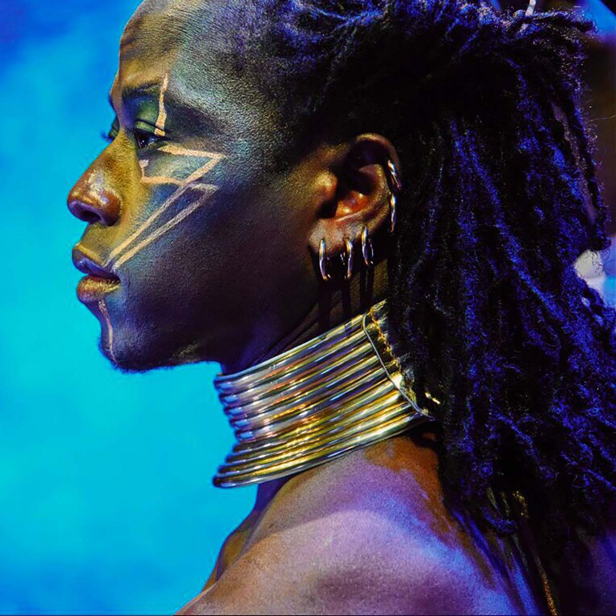 A member of Cirque Kalabante with gold rings around his neck, pierced ear and a design drawn on his face, staring intently to the side in this profile