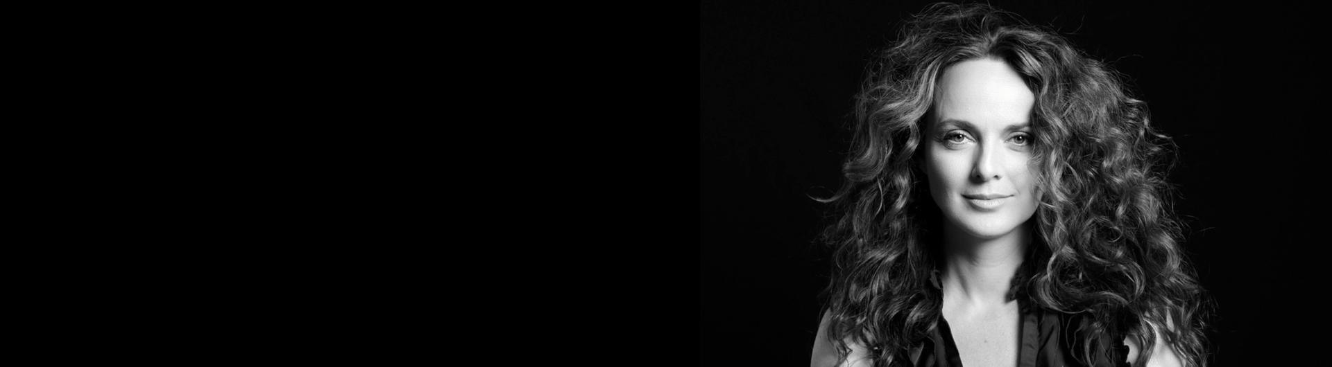 Melissa Errico with long curly hair gazes out at the viewer against a dark background