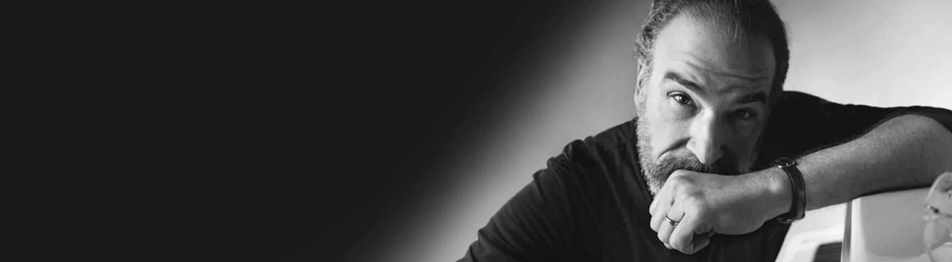 Black and white photo of Mandy Patinkin resting his arm on a piano.