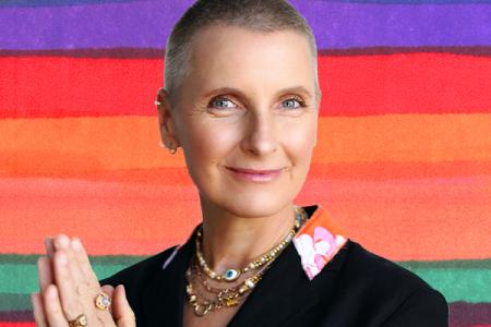Elizabeth Gilbert with her hands held together, smiling gently in front of a colorfully striped background.