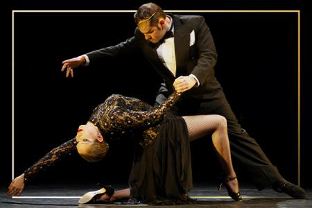 Two tango dancers in formal attire dancing, the woman swept back and held off the flower by the man who has his other arm outstretched.